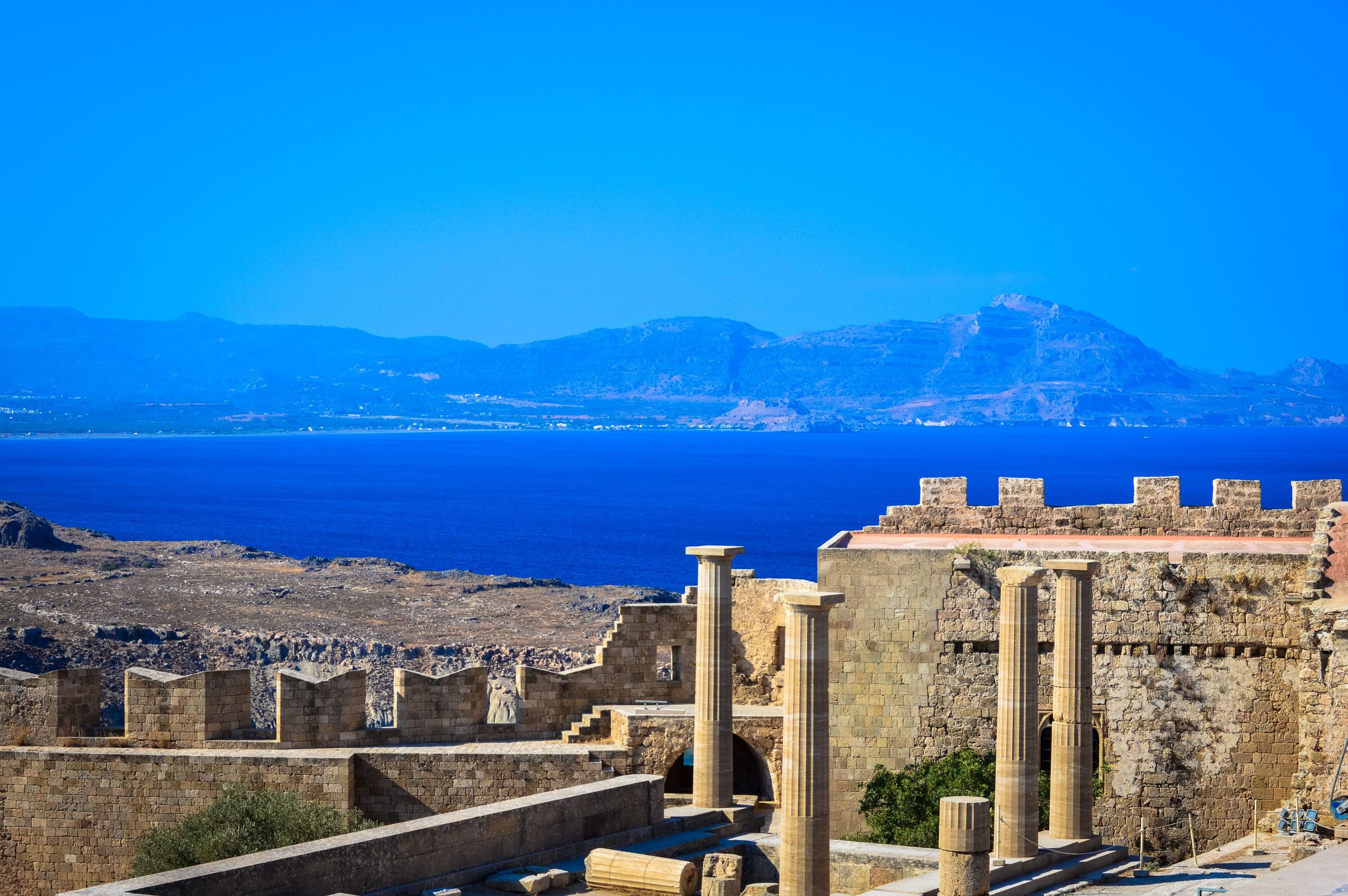Blog Scenic view of ancient Greek ruins with stone columns and walls against a vibrant blue Aegean Sea backdrop.
